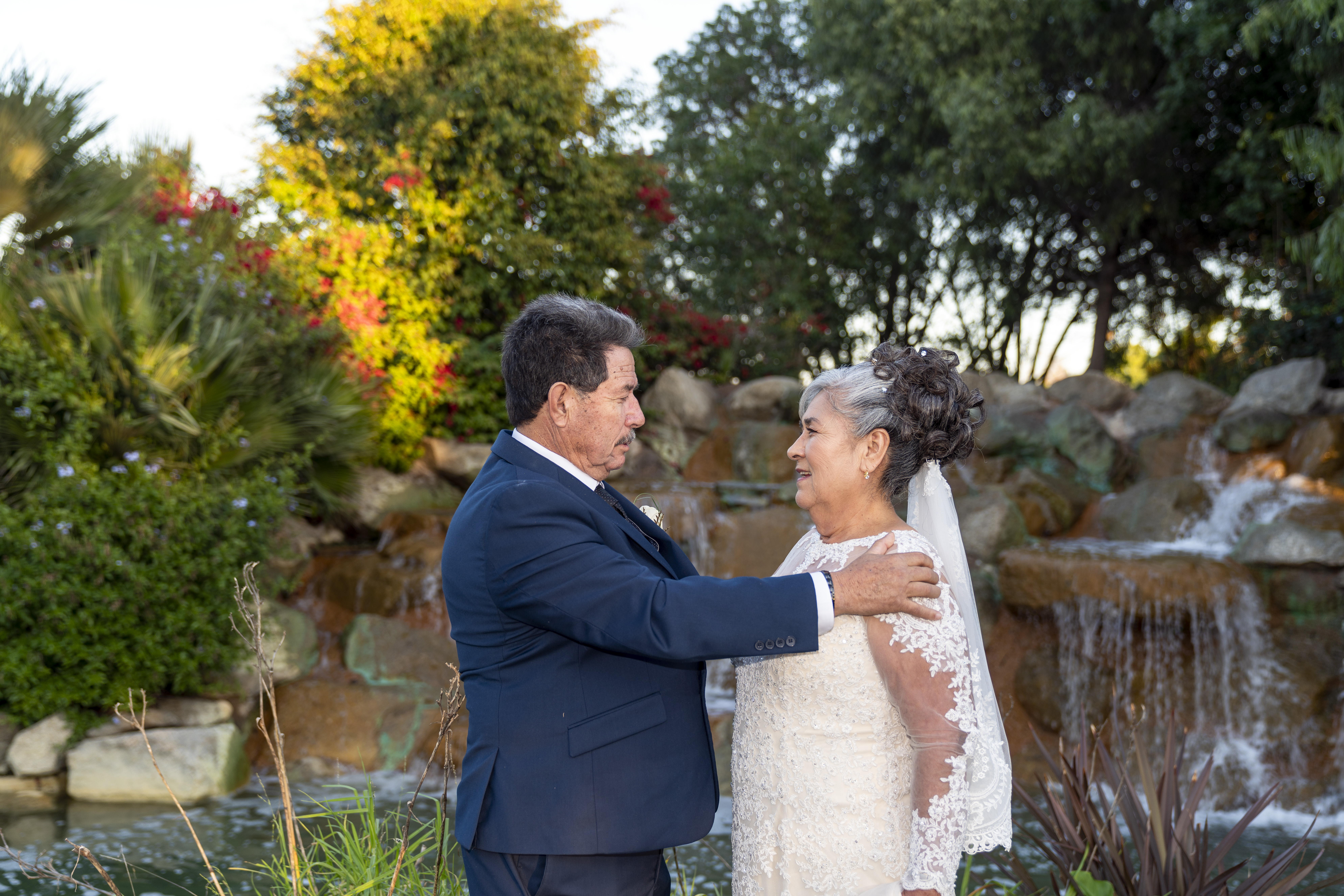 Couple celebrating their fiftieth anniversary, embracing and looking at each other beside a garden waterfall