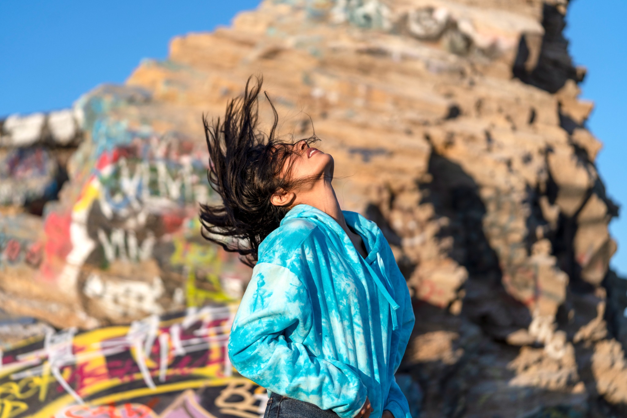 Danielle in a turquoise hoodie tossing her hair in sunlight against painted coastal ruins