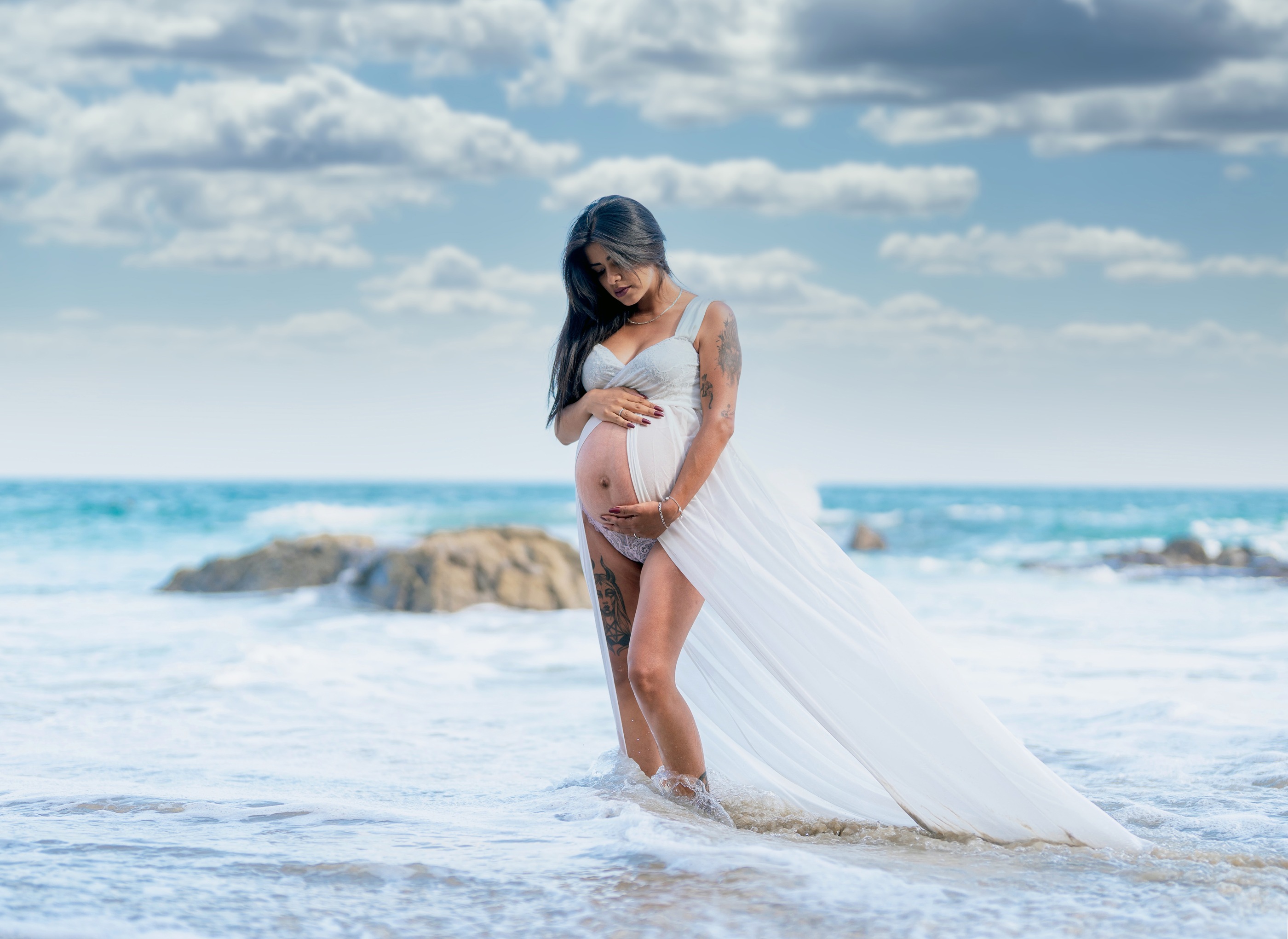 Maternity portrait in ocean surf, with a flowing white dress beneath a cloud-filled sky
