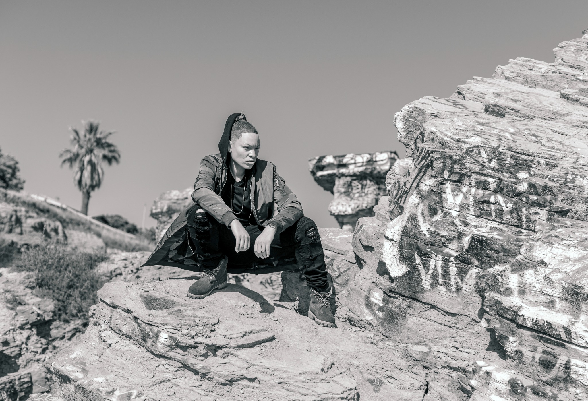 Black and white portrait of Shaunta seated on rocks among painted coastal ruins, shown from head to boots