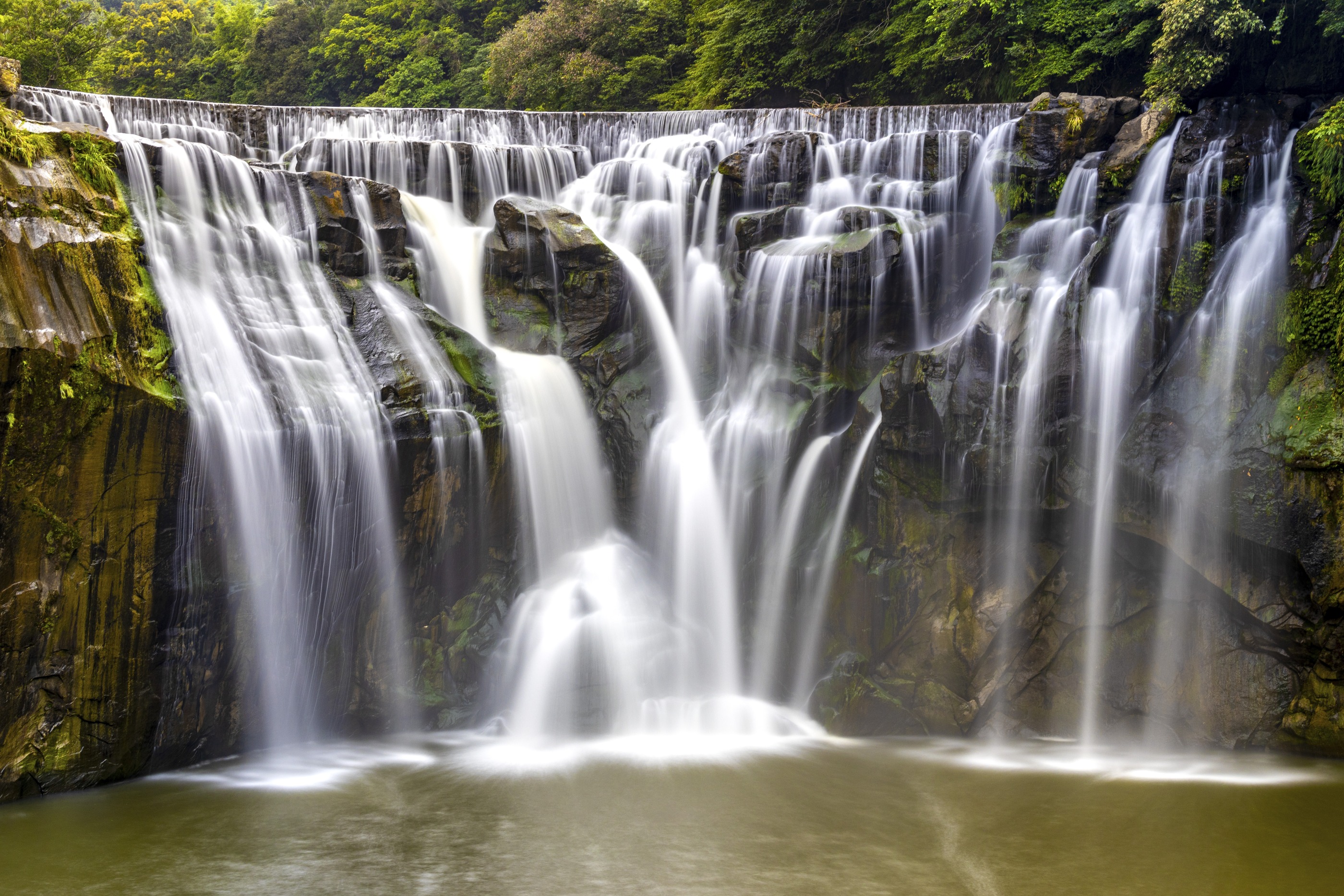 Silky streams of water cascading over a broad rocky waterfall surrounded by lush green forest
